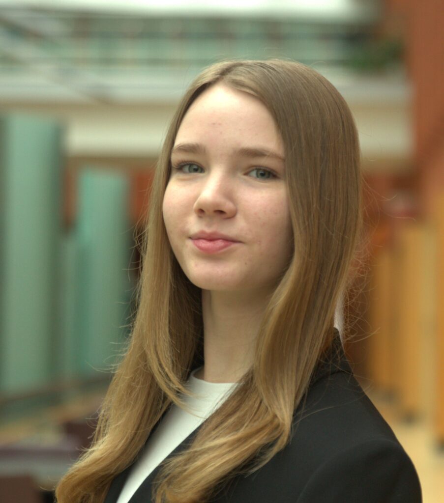 Kyra Sherman, a young woman with long, straight blonde hair, stands confidently in a softly lit hallway wearing a black blazer and white top, gazing directly at the camera.