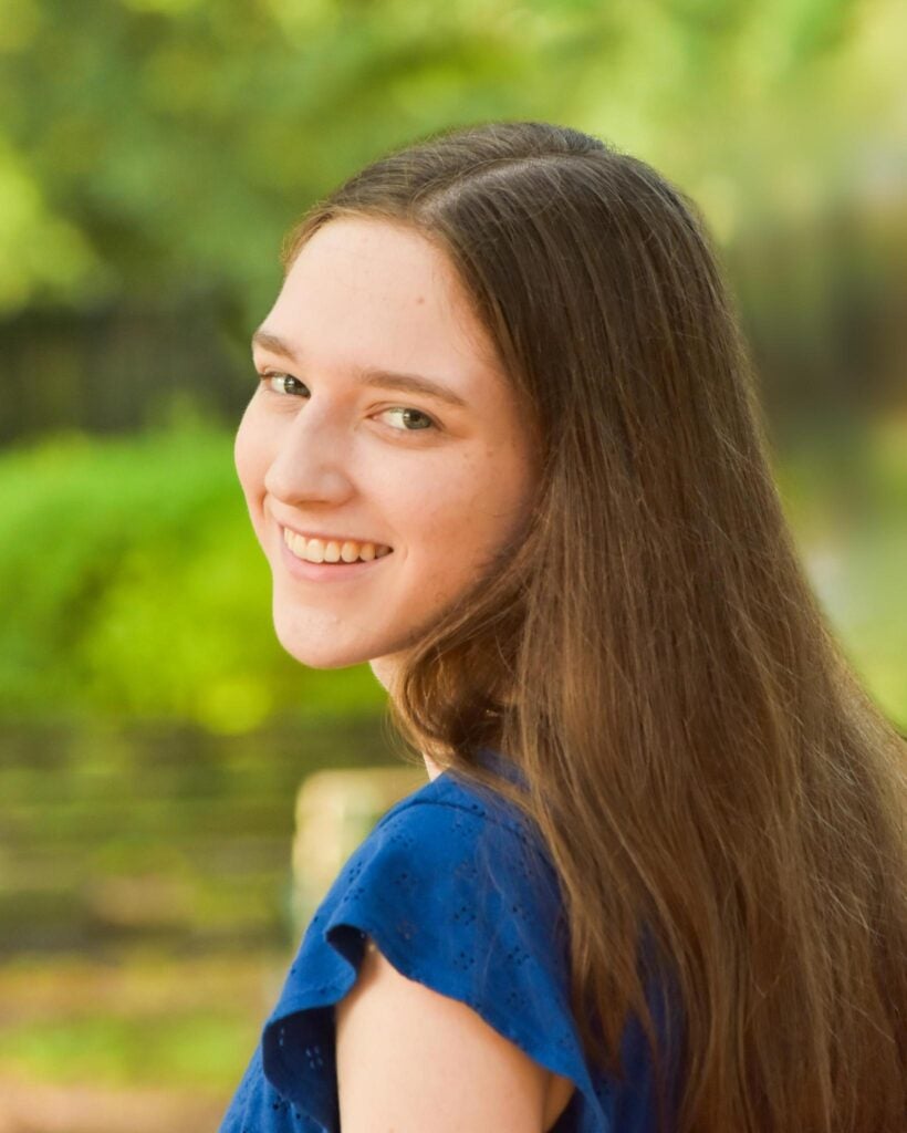 A young woman with long brown hair and a blue top smiles over her shoulder, standing outdoors with a blurred green background.