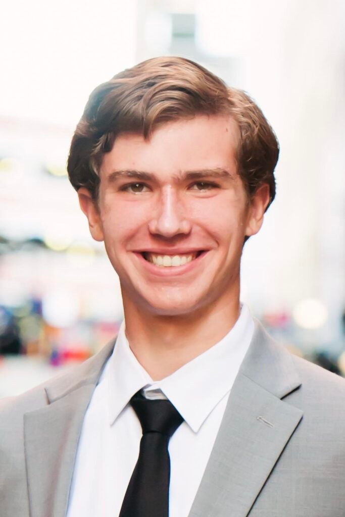 Charles Baker, a young man in a light gray suit, white shirt, and black tie, smiles at the camera outdoors with a blurred city street in the background.