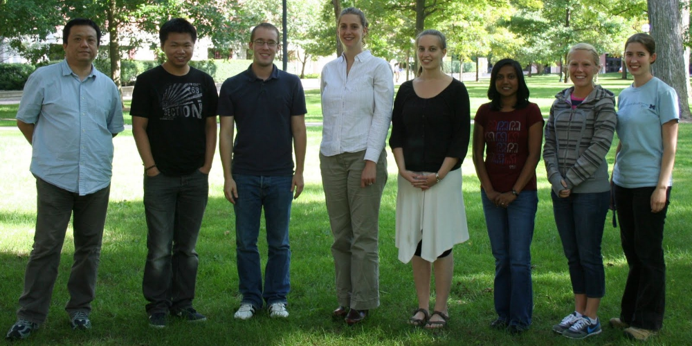 Seven people stand in a row on grass in a park, with trees and sunlight in the background. They are facing the camera and smiling, dressed in casual clothing.
