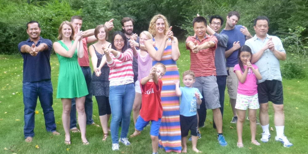 Thirteen adults and two children stand on grass, smiling and posing with playful hand gestures in a wooded outdoor area.