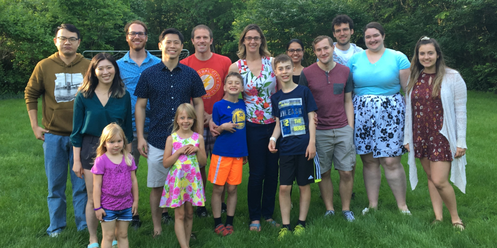 A group of sixteen people, including adults and children, stand smiling on a green lawn with trees in the background. Some wear casual summer clothing, and two young girls stand in front.