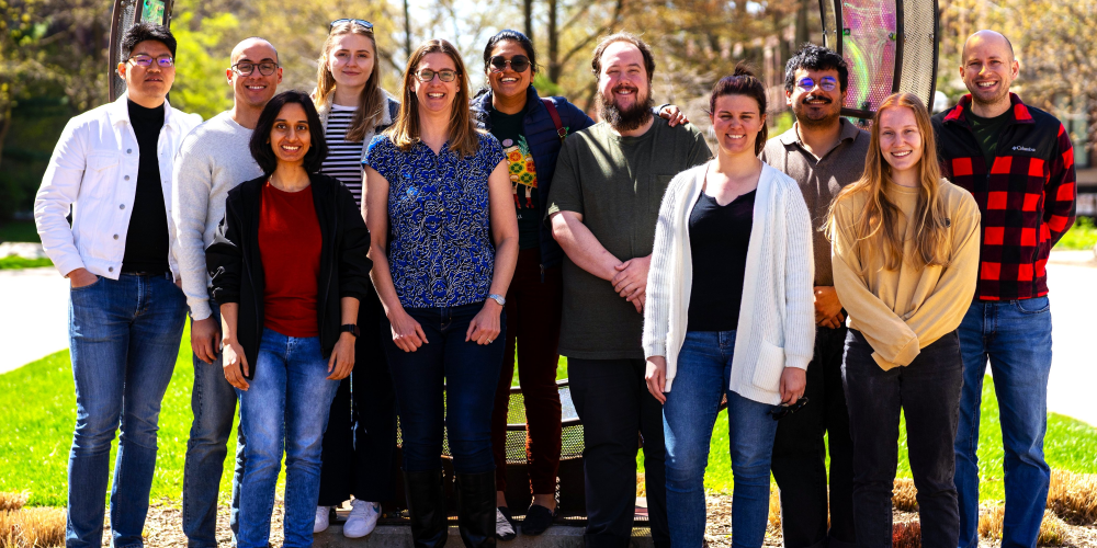 A group of eleven people stand outdoors in bright sunlight, smiling at the camera. They are dressed casually, with trees and green grass visible in the background.