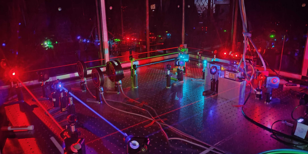 A dark laboratory setup with multiple red and blue lasers intersecting across a metal optical table, mirrors, lenses, and scientific equipment reflecting colorful lights.