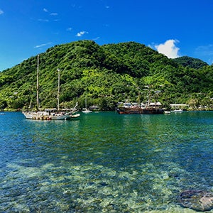 Image of coastal region in Pago Pago on Tutuila, American Samoa. Several boats moored in a lagoon along the American Samoa coastline with forested mountains in the background.