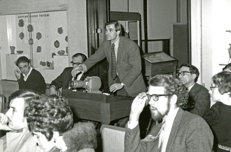 man in suit handles a slide projector while seated audience members look on.