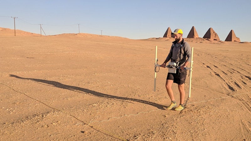 Man walking in desert with scientific equipment, pyramids in background