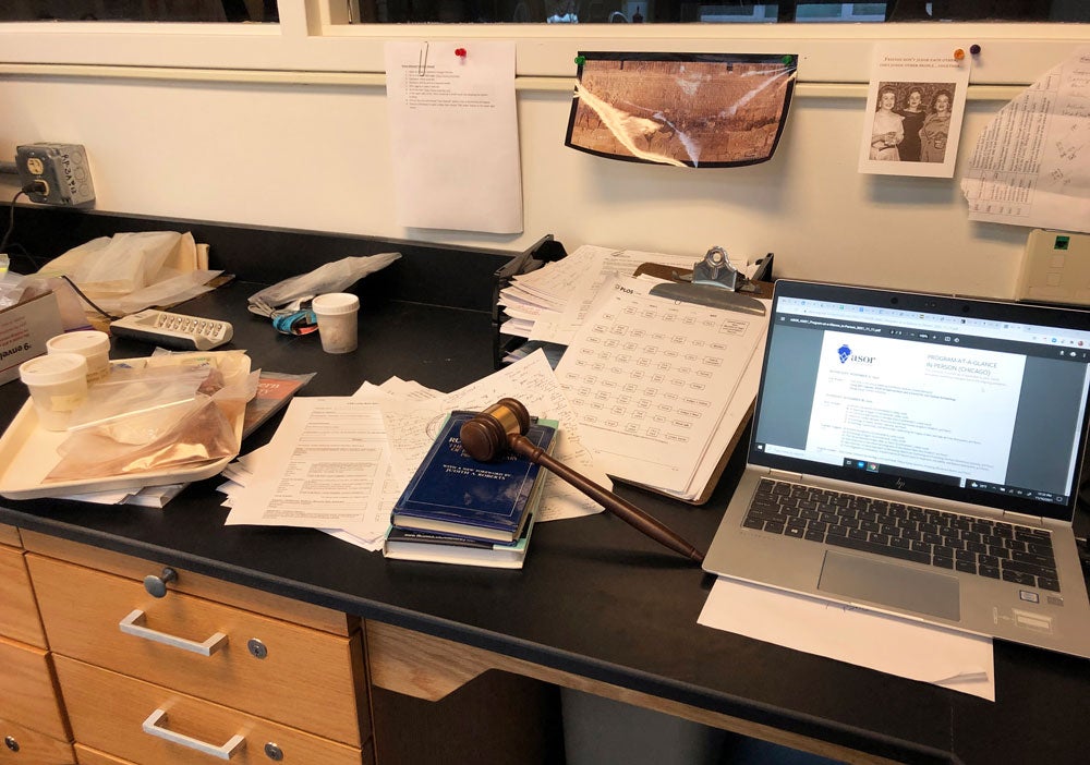 color photo of a desk strewn with papers and an open laptop. A gavel rests on top of a short stack of books.
