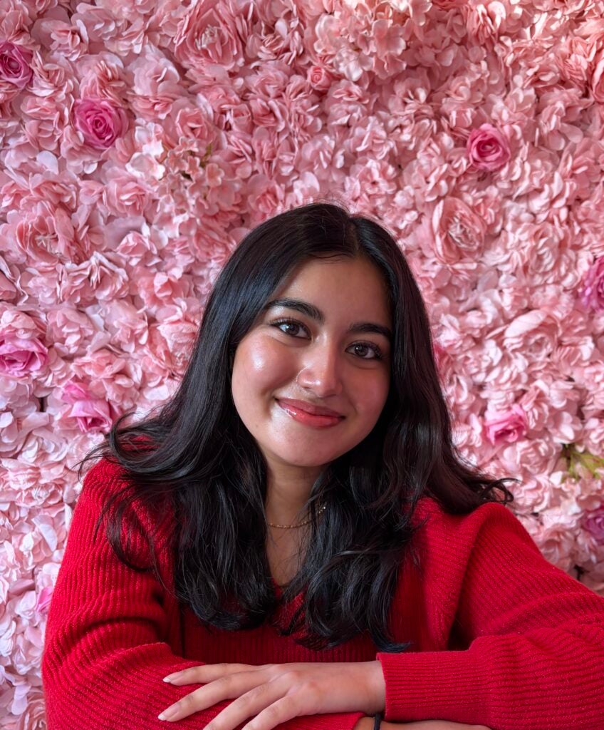 A woman with long dark hair and a red sweater smiles in front of a wall covered with pink and light pink flowers.