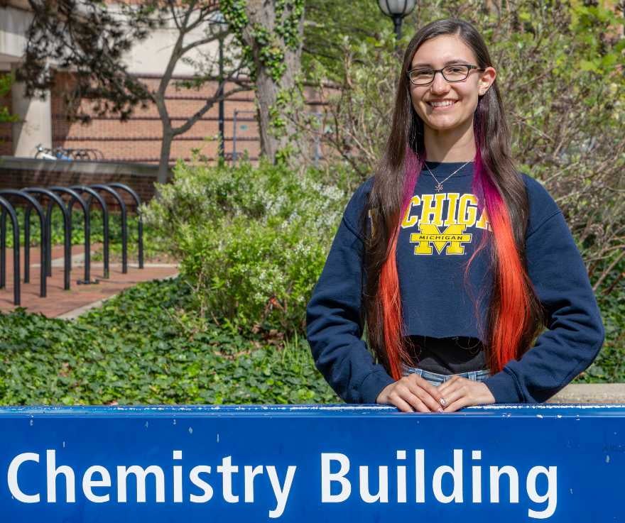 A person with long brown and red hair, glasses, and a Michigan sweatshirt stands smiling behind a blue sign that reads Chemistry Building, with greenery and a bike rack in the background.