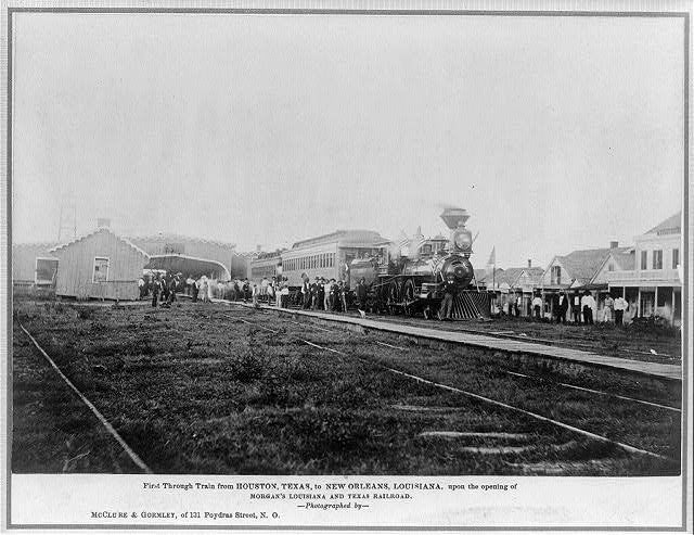 Historic black-and-white photo of a steam locomotive and passenger cars at a train station, surrounded by a crowd of people. Early 1900s-style buildings line both sides of the tracks.