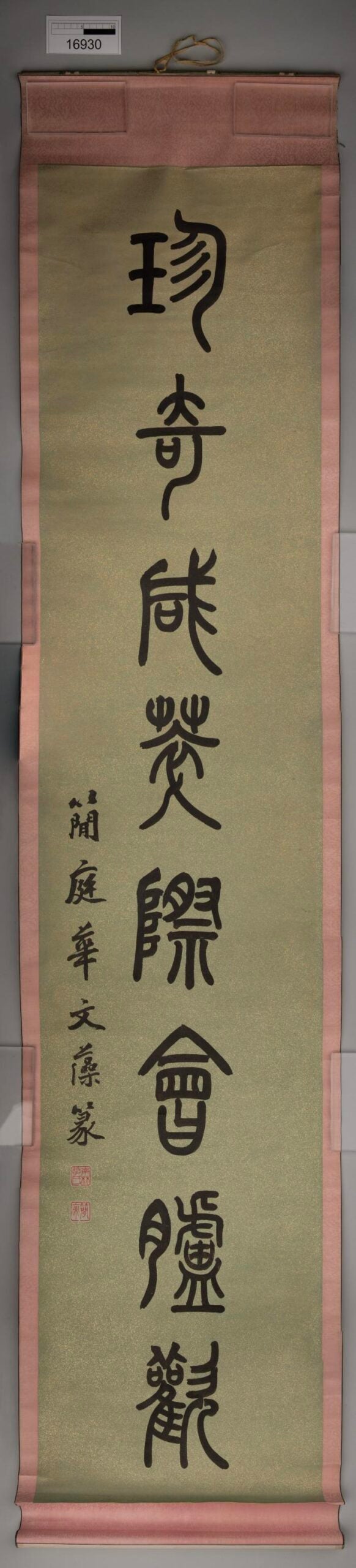 A vertical hanging scroll with Chinese calligraphy in bold black ink on a light tan background, bordered with pink fabric. Four large characters are arranged top to bottom, with smaller text and red seals at the bottom.
