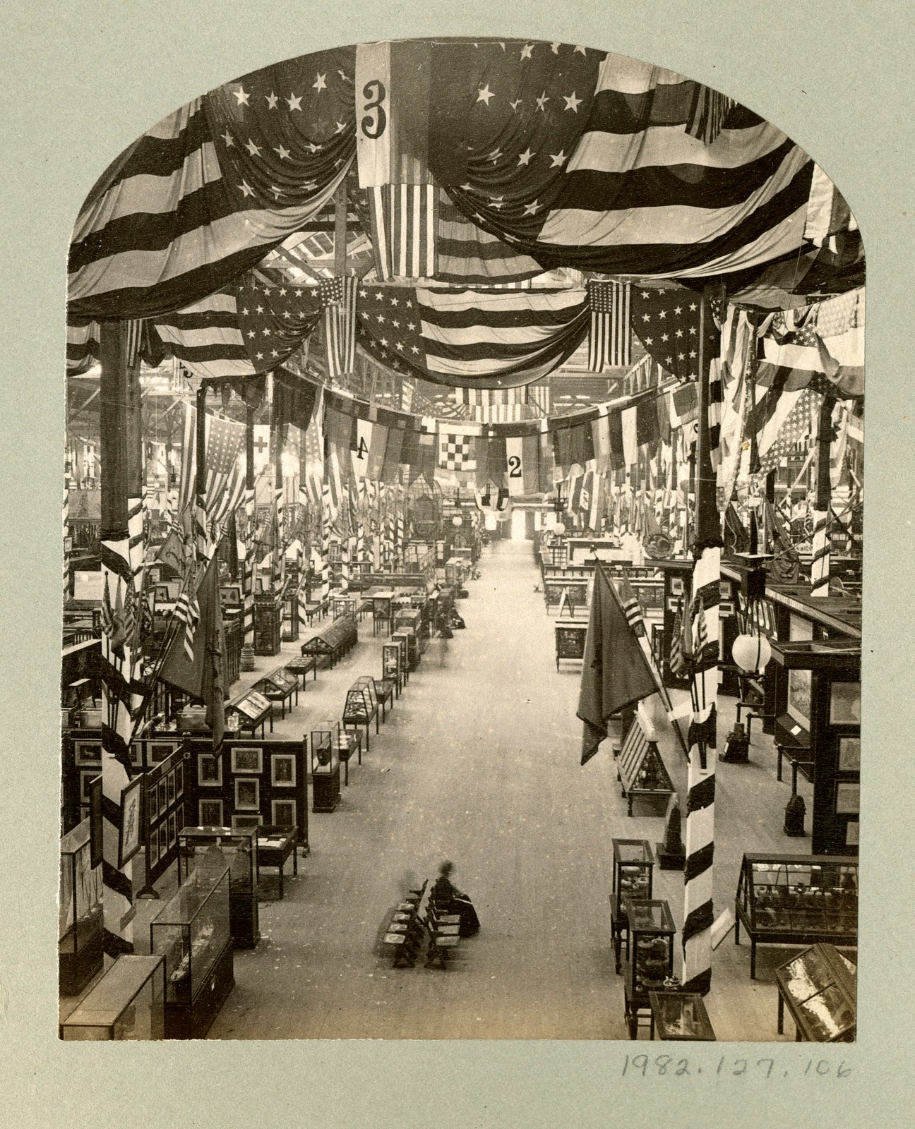 Large exhibition hall decorated with American flags and draped fabric. Display cases and framed pictures line the wide central aisle. Numbered signs hang from the ceiling above exhibition areas.