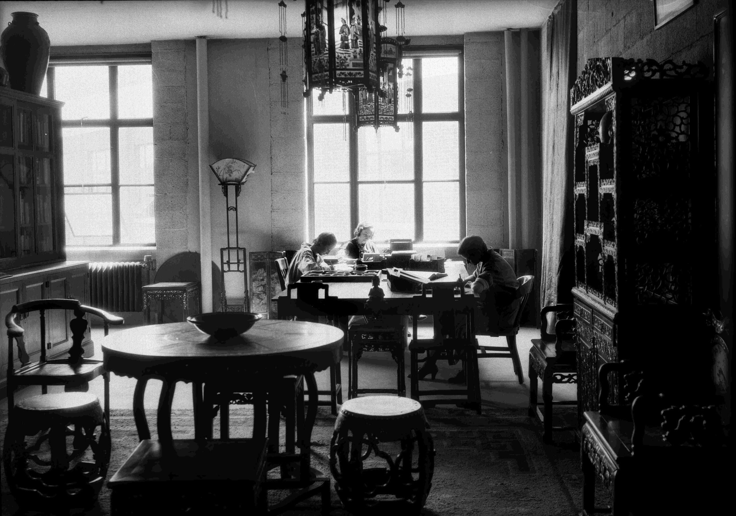 Two people sit and work at a table in a sunlit room filled with ornate, traditional Chinese furniture and decorations, including carved chairs, screens, and a hanging lantern. Light streams in through large windows behind them.