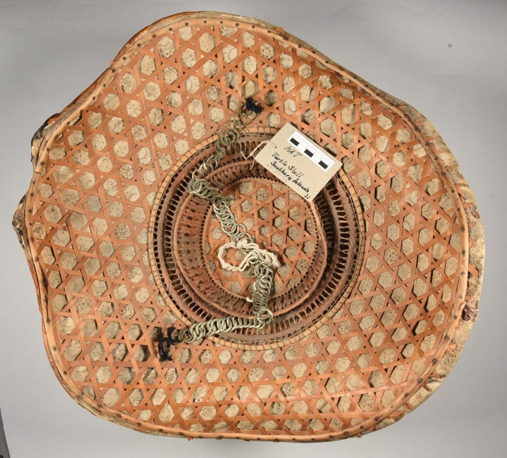 The bottom side of the turtle shell salakot. It has rattan weaving on it, along with a chain neckstrap attached with dark string. The tag attached to the hat reads "HAT; Turtle Shell; Southern Islands; 21"