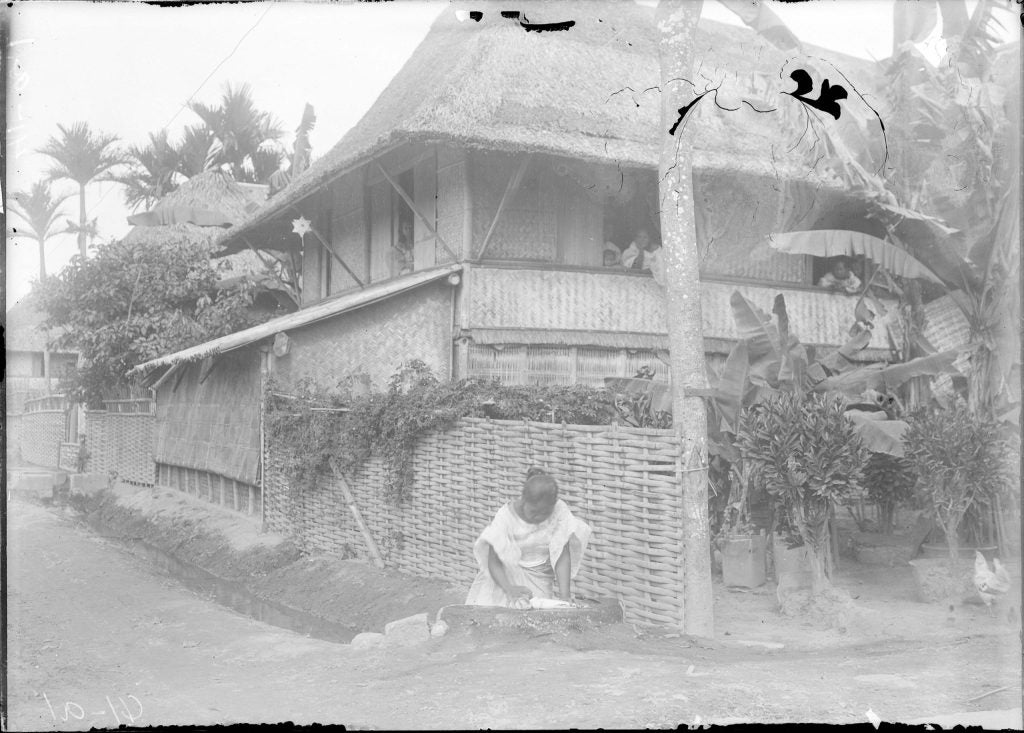 An image of a young woman scaling fish outside of a house. In the house, several more young women are looking out from three different windows.
