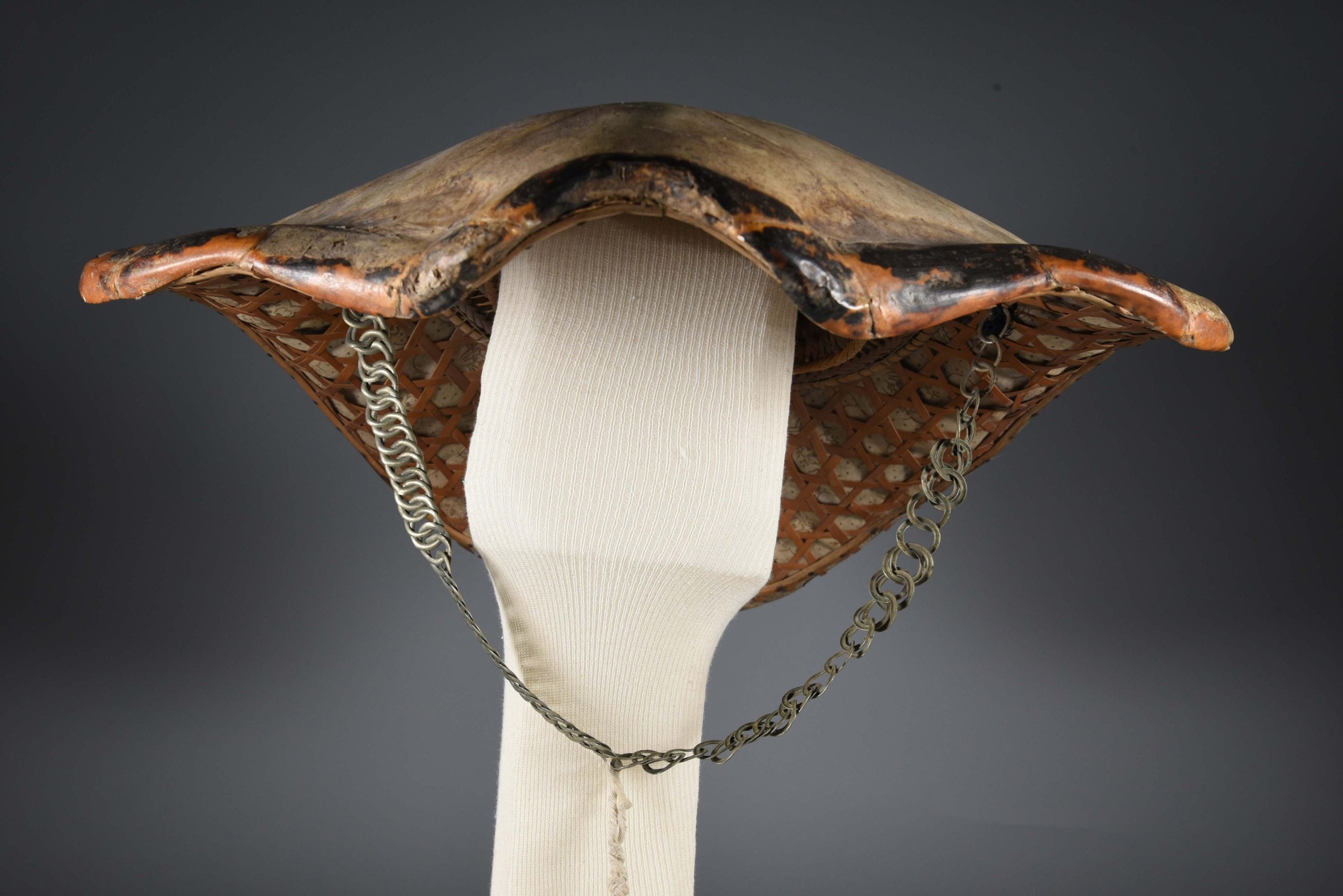 An image of a turtle shell hat, or salakot, sitting on top of a mannequin head. From this view, the shell takes on the appearance of a swimming turtle, with the chain neckstrap hanging underneath.