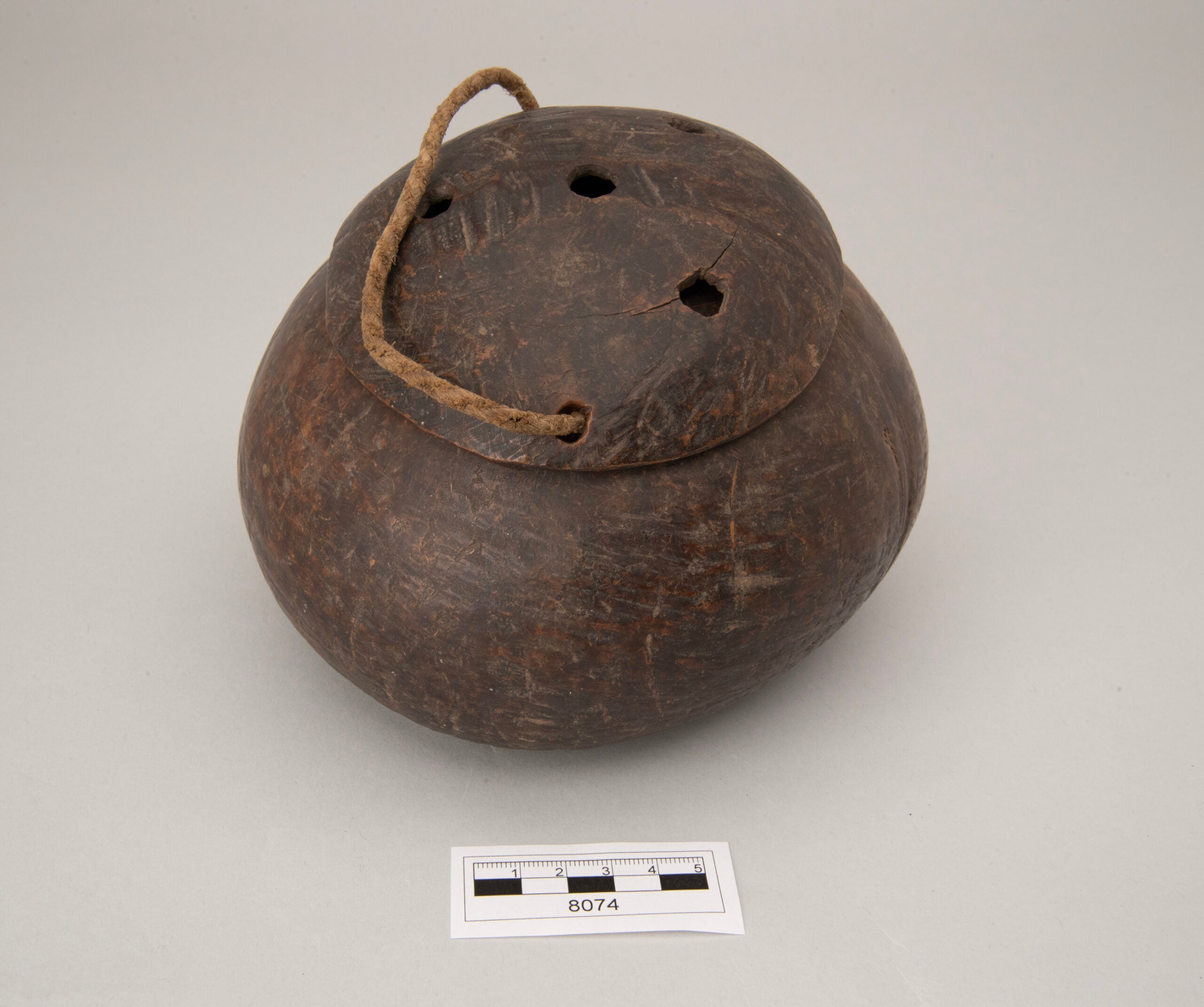 A container made out of out a coconut shell, with its lid on. Three holes on the top of the lid and an attached string are visible.