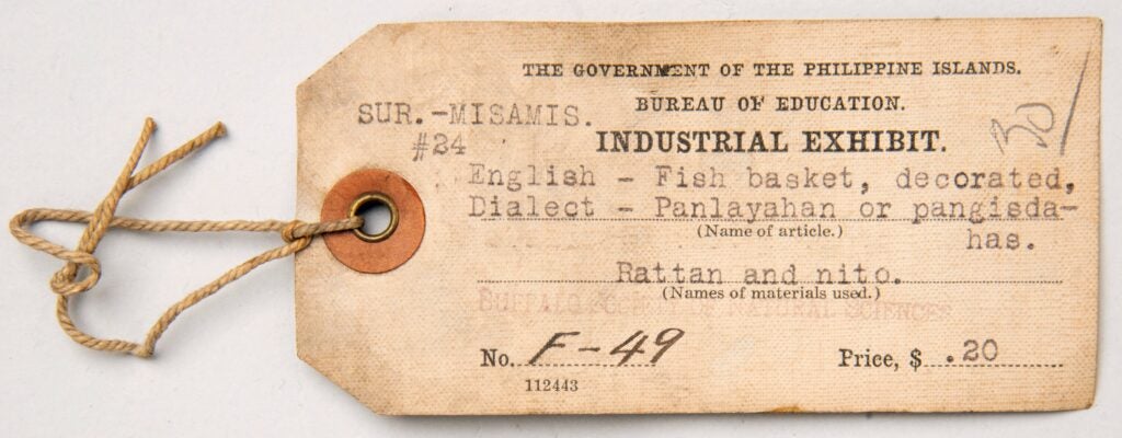 Image of rectangular cloth label yellowed with age. The label title is printed in black all caps serif font, “The Government of the Philippine Islands. Bureau of Education. Industrial Exhibit.” A stamped faded and discolored with aged is barely legible that reads, "Buffalo Society of Natural Sciences".
