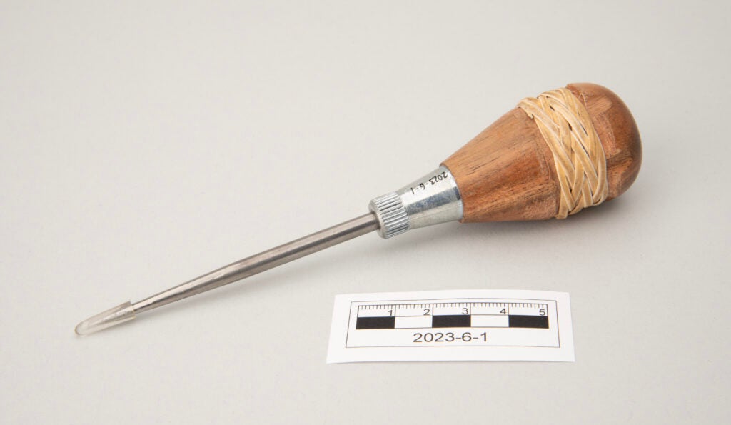 This image shows a pointed metal awl used for bamboo weaving. The tool has a long silver metal shaft ending in a sharp point protected by a removable plastic cover. The rounded wooden handle is wrapped with woven rattan. The handle appears light brown with visible wood grain and natural fiber texture.