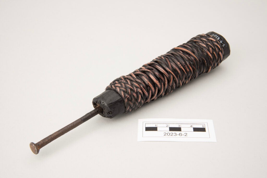 This image shows a bamboo weaving tool consisting of a flat-head iron nail attached to a wooden handle. The handle is cylindrical and appears dark brown to nearly black, possibly lacquered or painted. A woven rattan covering wraps most of the handle, with traces of the original light tan rattan visible through areas of wear. The iron nail extends from one end of the handle and likely served as a tool for manipulating or separating bamboo strips during weaving.