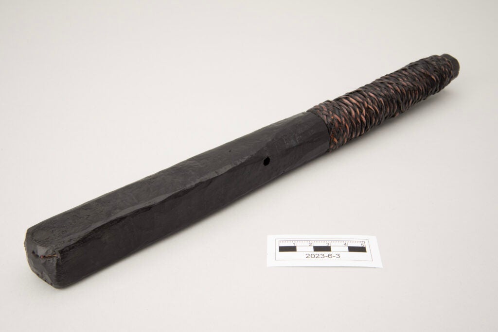 This image shows a flat, rectangular wooden bat used to flatten bamboo for weaving. The bat appears to be lacquered or painted a glossy dark brown, nearly black. The handle is wrapped with woven rattan, also coated in the same dark finish, with traces of the original light tan rattan visible through areas of wear.