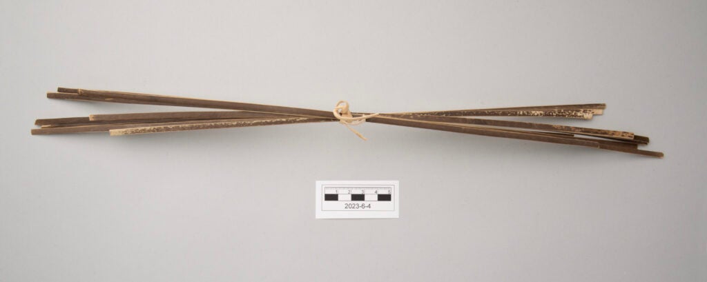 This image shows a bundle of split bamboo prepared for weaving. The bundle contains eleven bamboo strips of similar size tied together with a thin piece of pale bamboo. Each strip shows two distinct surfaces, with one side dark brown and the other pale tan. The strips curve slightly inward, with the lighter tan surfaces facing the interior of the bundle.