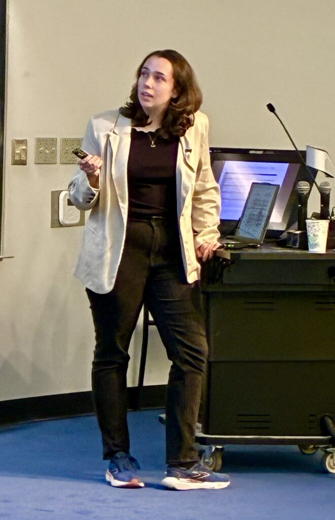 A woman in a light blazer stands next to a podium with a laptop and microphones, holding a remote and presenting her summer research with grace in a classroom or lecture hall.