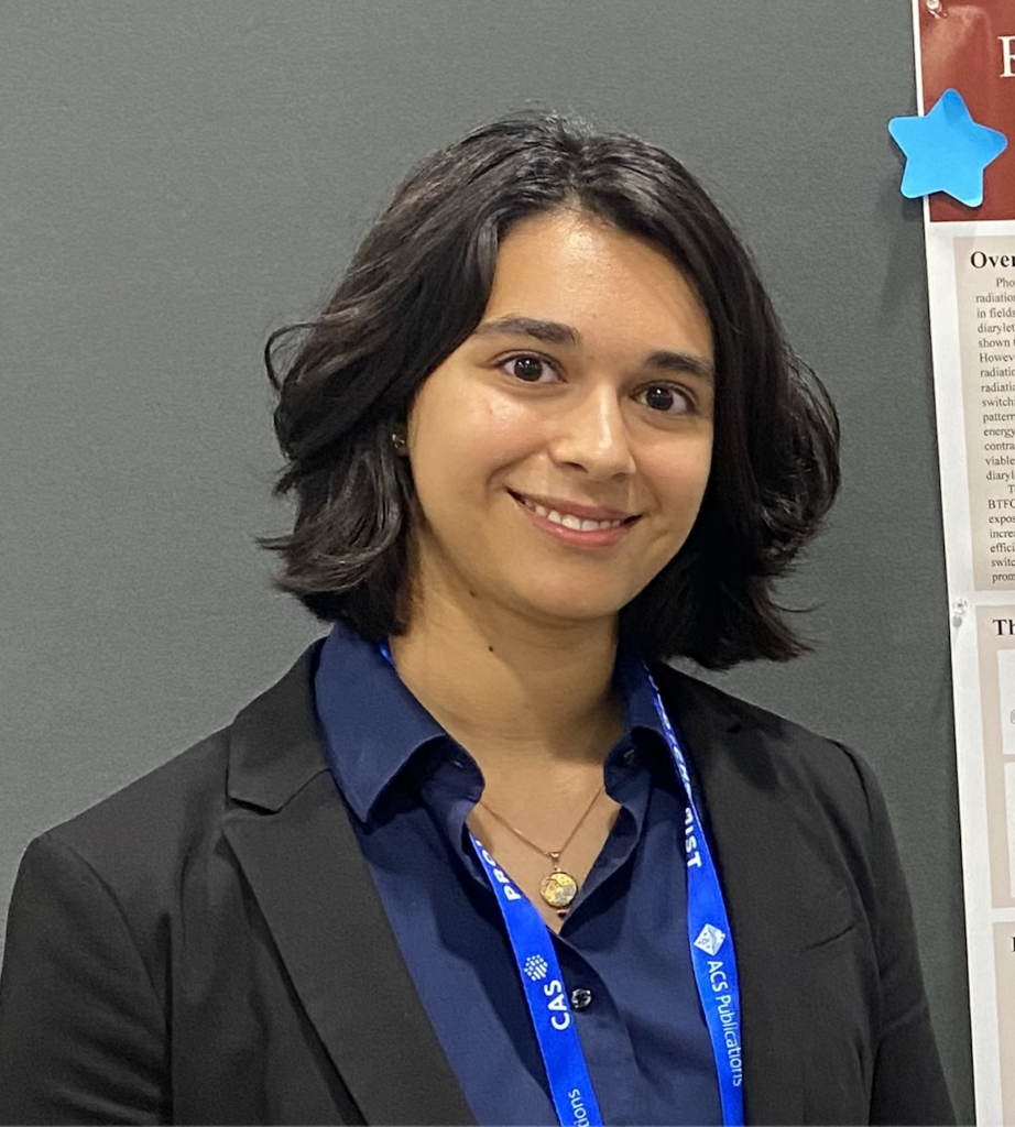 A person with short dark hair, wearing a navy shirt, black blazer, and a blue lanyard, smiles in front of a gray wall and a poster with a blue star sticker.