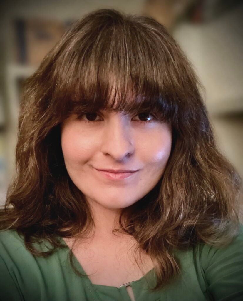 A person with wavy brown hair and bangs smiles softly at the camera, wearing a green top. The background is softly blurred.