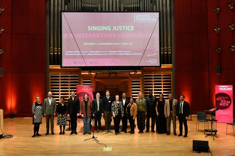 15 individuals pose on a stage with bright pink banners and screen projection for the Singing Justice conference