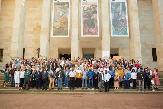 A large group of people wearing conference nametags pose on steps in front of a building with tall square columns and three large banners of artwork.