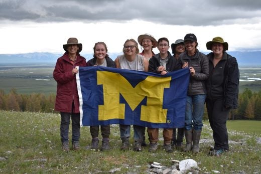 Eight individuals smile and hold up a University of Michigan "Block M" flag; steppe and mountains in the distance.