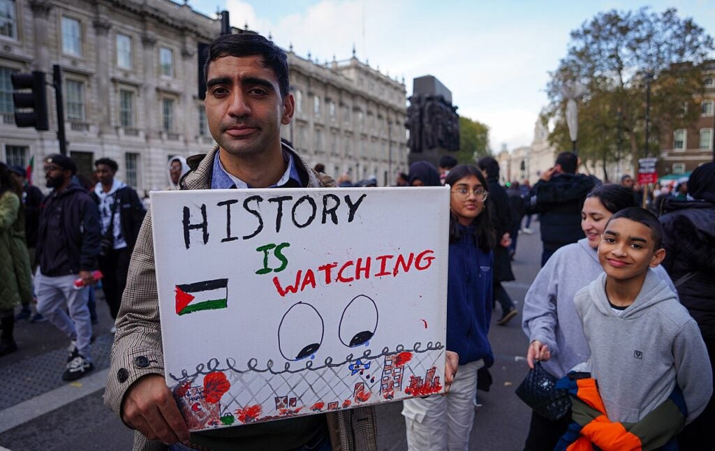 A man at a public demonstration holds a sign reading HISTORY IS WATCHING with a Palestinian flag and illustrated eyes, highlighting the semicivilized tactics faced. Several people, including children, stand around him on a city street.