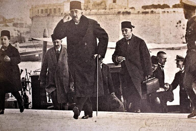 A group of men in early 20th-century attire, displaying Ottoman heuristics, walk away from a docked boat. One man in front uses a cane and raises his hand to his head, while others follow him. A large stone building is in the background.