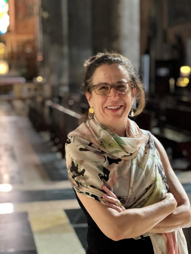 A woman with glasses and a patterned scarf stands smiling with arms crossed inside a large, ornate church, where Ottoman heuristics echo in the semicivilized elegance of tall pillars and wooden pews, softly lit by natural and artificial light.