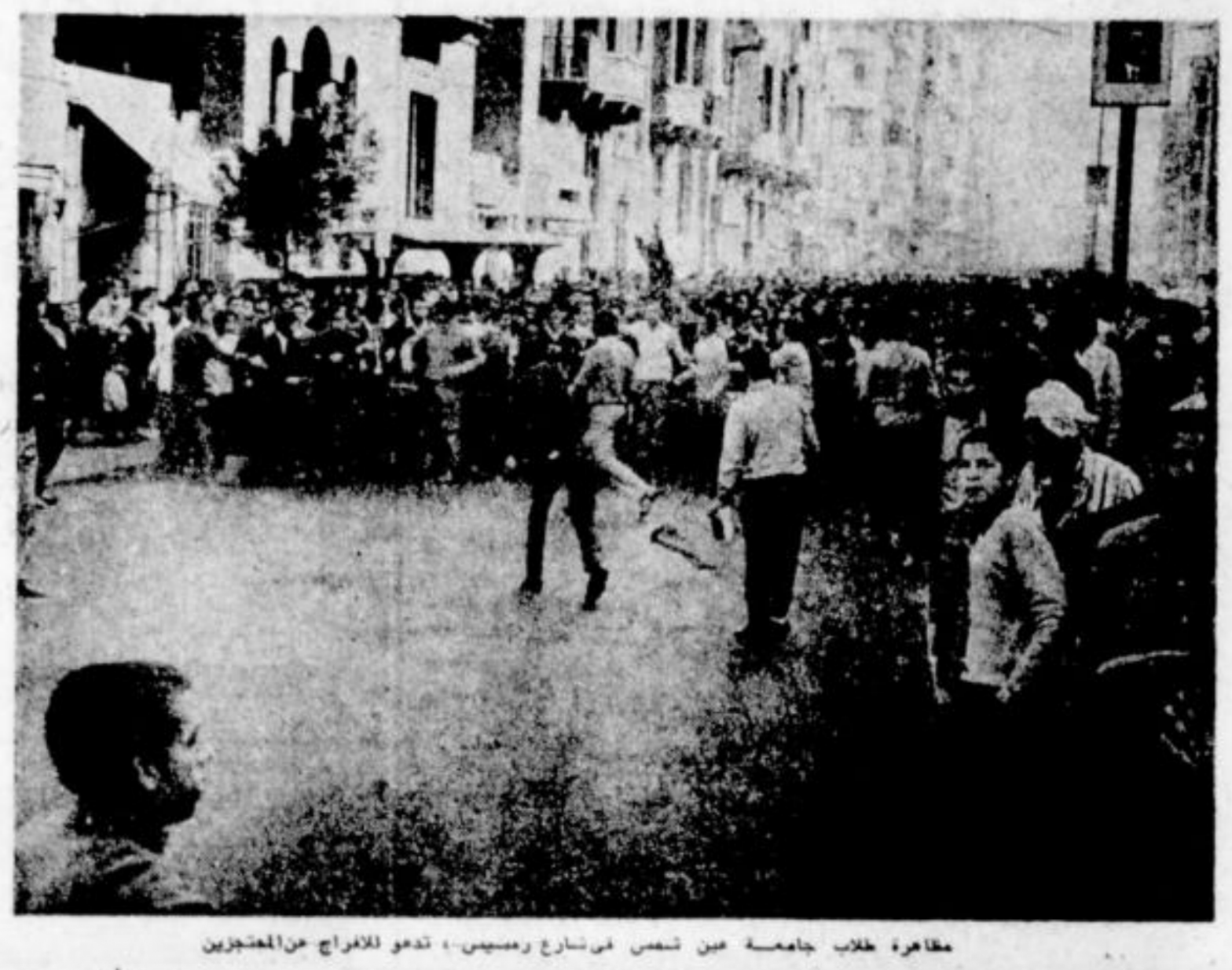 A large crowd gathers on a city street, some appearing to protest while others observe. The busy scene and surrounding buildings evoke an urban environment. The black and white image recalls moments from protest history, such as the Egyptian protest.