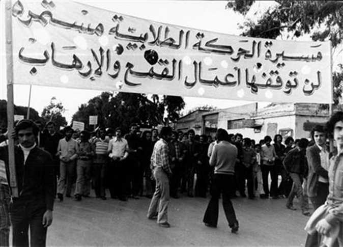 A black and white photo shows a group of people gathered under a large banner in Arabic that reads: The student movement march continues, it will not be stopped by acts of repression and terrorism—a striking moment in Egyptian protest history. Trees and buildings are in the background.