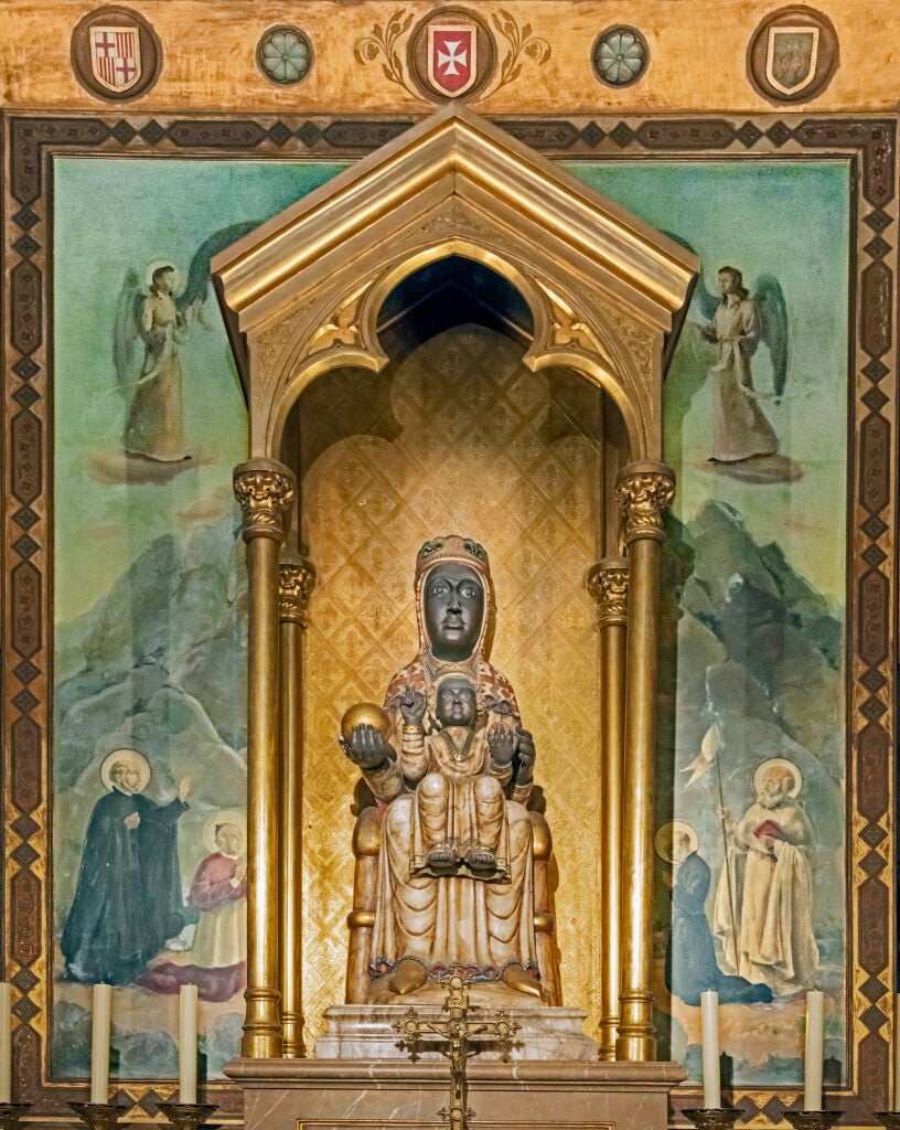 A black Madonna and Child statue sits beneath a golden canopy, surrounded by gold columns. The backdrop features painted angels and saints against a green and gold background, celebrating faith with prominent religious emblems above.