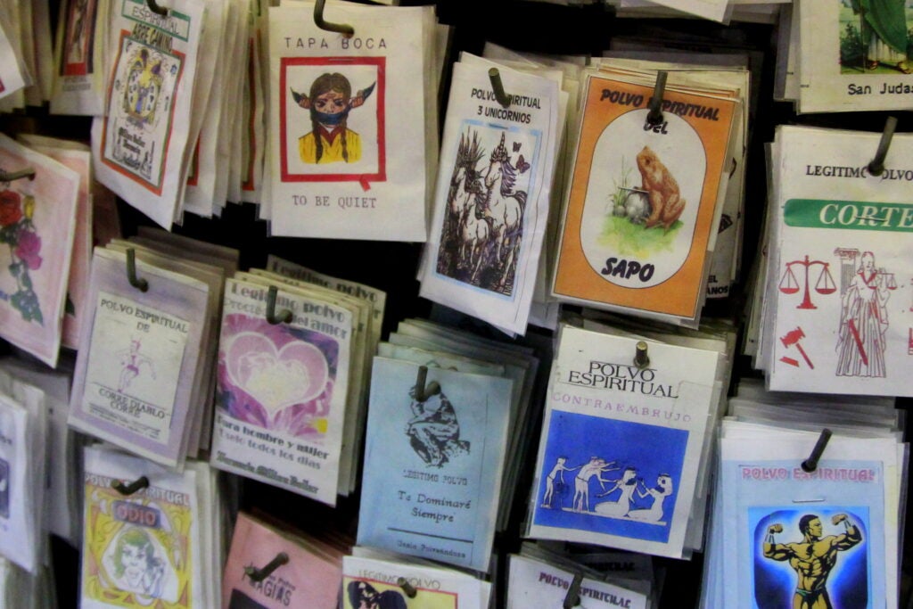 Packets of spiritual powders with colorful illustrated labels in Spanish, featuring various animals, symbols, and mystical imagery tied to faith and materiality, are displayed hanging on hooks for sale.