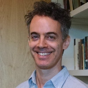 A man with short, curly dark hair and a light blue collared shirt smiles at the camera. He stands indoors before shelves of books and a wooden door, reflecting on religious cooperation inspired by places like Mecca and the Shrine of Santa Rosalia.