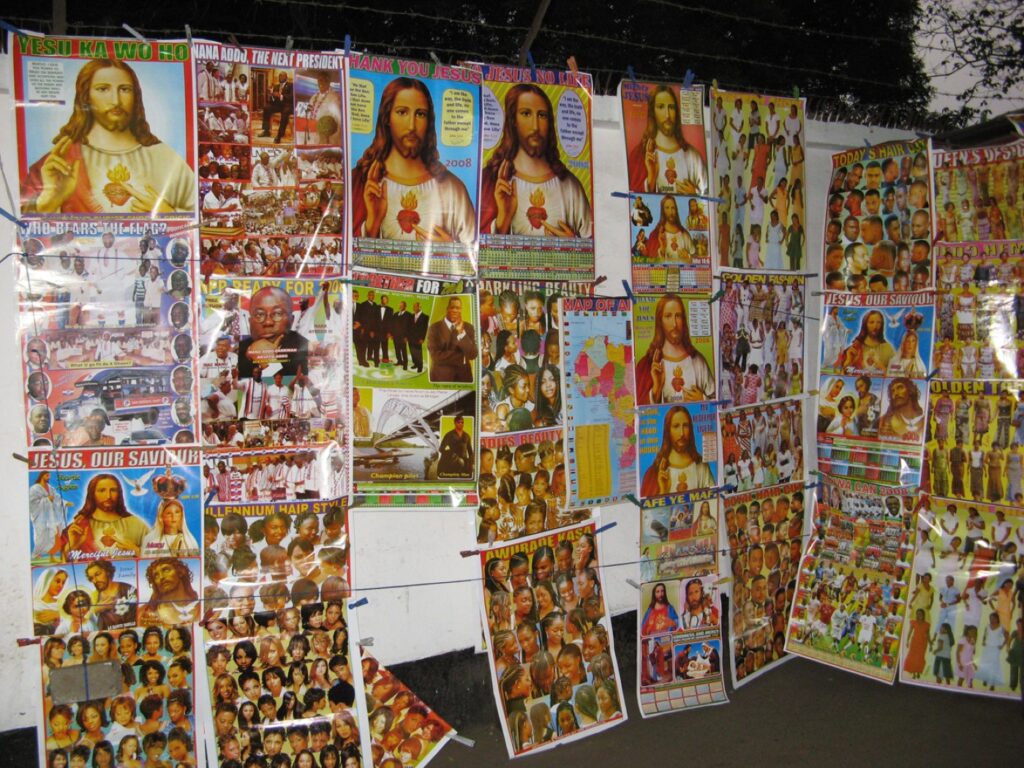 Colorful posters featuring various images of Jesus, messages of faith, and pictures of children are displayed on a wall outdoors, arranged in overlapping rows and columns.
