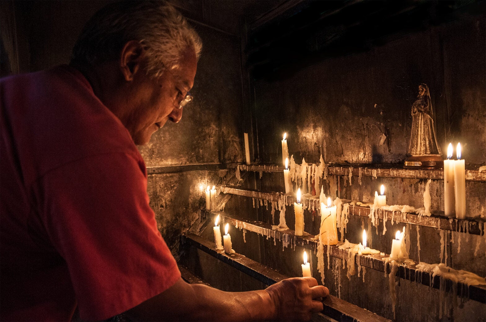 An older man in a red shirt lights a candle at an altar with many burning candles and an image of the Virgin Mary, invoking a reflective atmosphere where materiality meets the quiet devotion of religion.