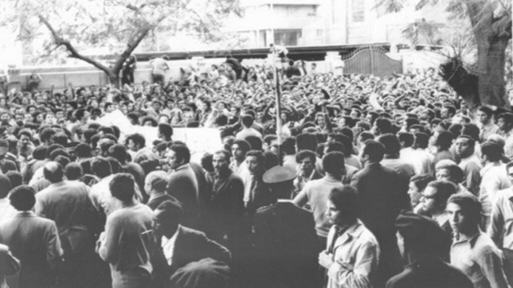 A large crowd of people, mostly men, gather outdoors in black and white for what appears to be an Egyptian protest. Some hold signs, and a few uniformed individuals are present among the crowd. Trees and buildings are visible in the background.