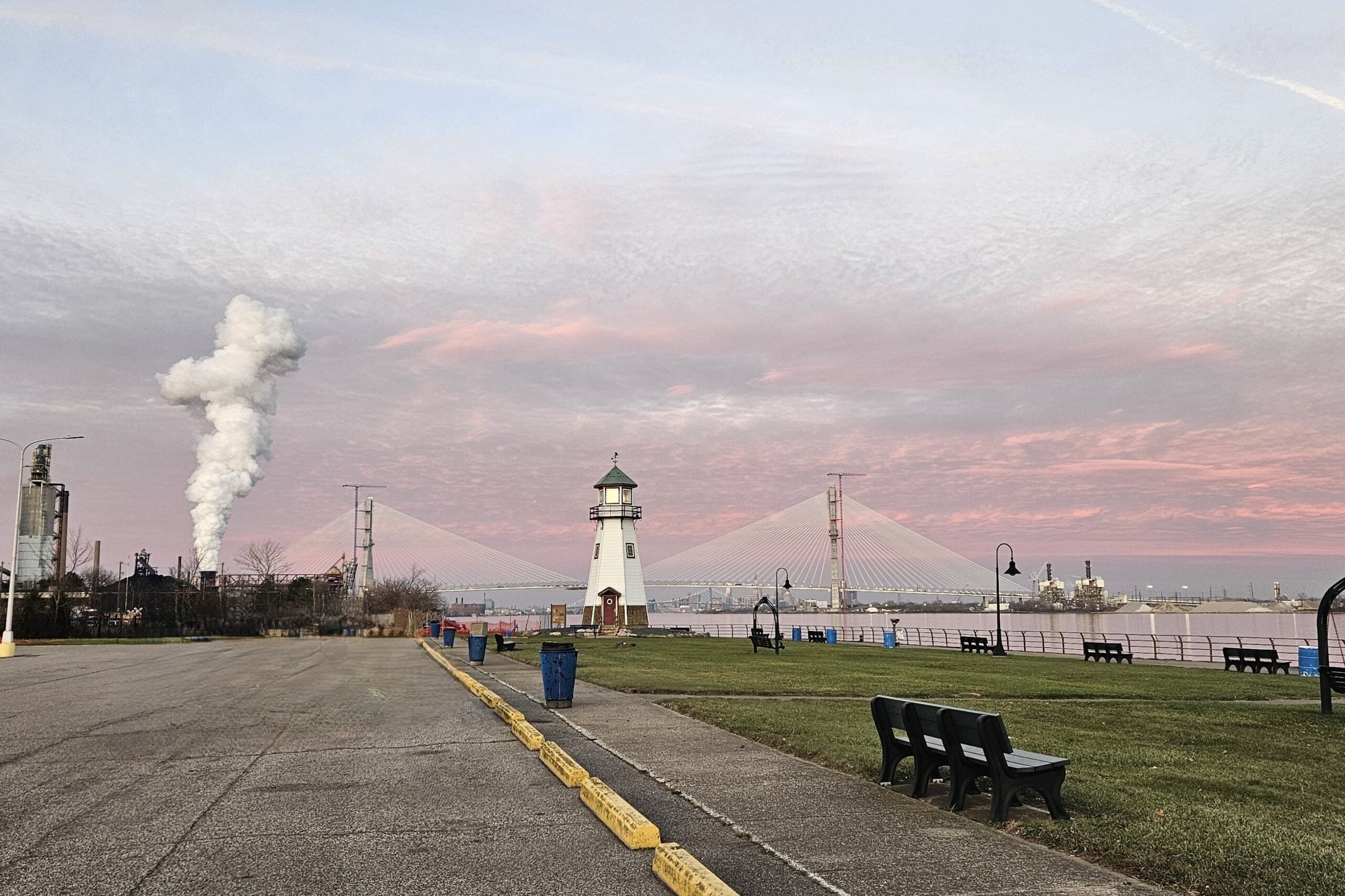 A lighthouse stands by a river with benches and trash cans nearby in Belanger Park. A suspension bridge and a factory with white smoke are in the background under a pinkish evening sky.