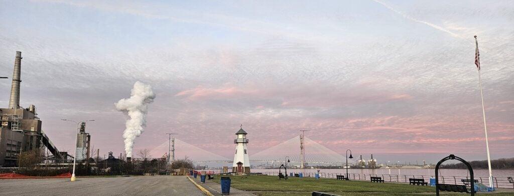 A riverside scene at sunset shows a lighthouse, a suspension bridge, industrial buildings with smoke, grassy parkland like Belanger Park, a flagpole, and a pink-hued sky—a perfect Field Trip spot highlighted in any Planning Guide.