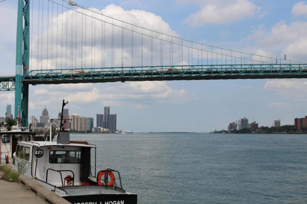 good-river-picture-1-scaled A boat docked by the river on the east side, with a large suspension bridge spanning overhead and the city skyline visible in the background under a partly cloudy sky.