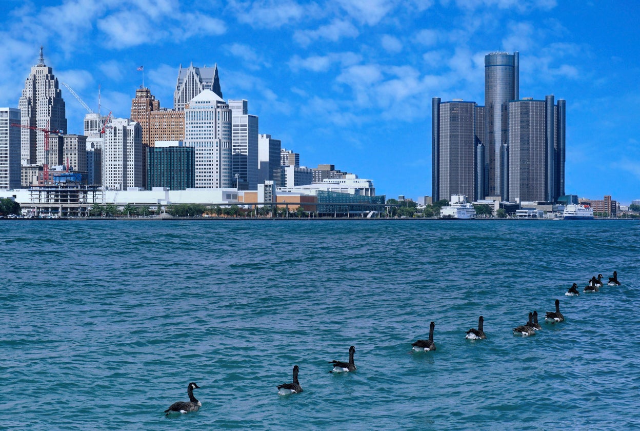 A line of geese swims across a blue river with the Detroit city skyline and tall buildings in the background under a bright blue sky with scattered clouds, not far from where the Detroit Port Authority works to cut emissions and promote sustainability along the waterfront.