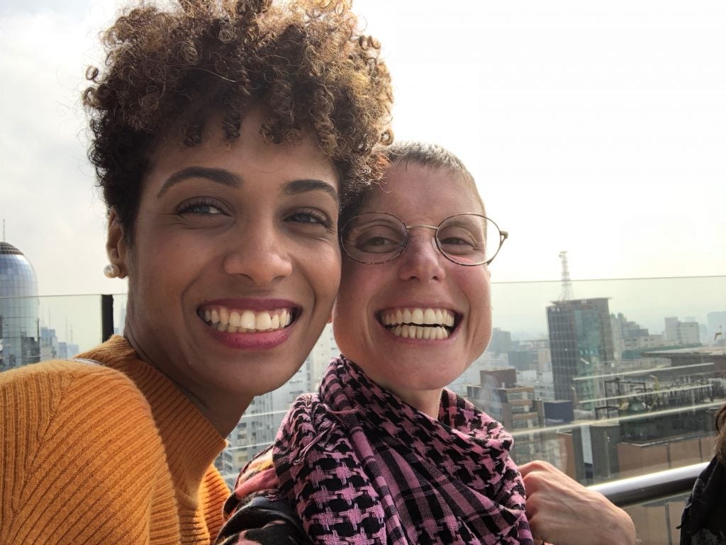 Stephanie Ribeiro and Marisol Fila smile at the camera as they take a selfie together with the city of São Paulo in the background during the summer of 2018