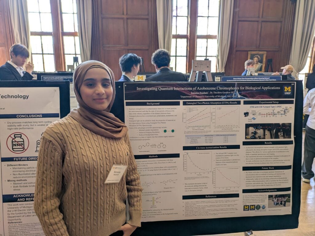 A young woman wearing a brown hijab and beige sweater stands in front of a scientific research poster at a conference. People and other posters are visible in the background in a large, well-lit room with tall windows.